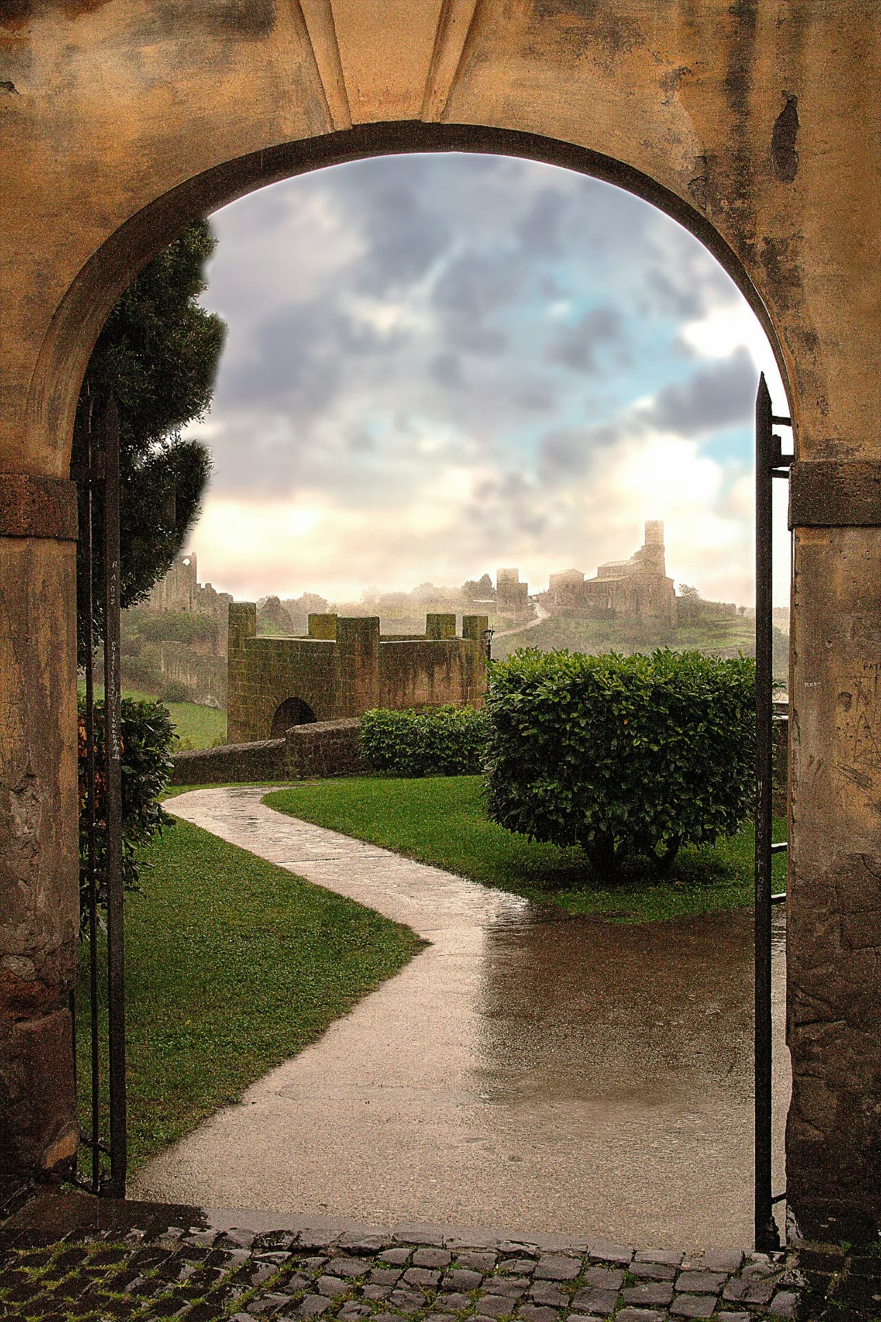 Through an arched gate in Tuscania, a wet path toward San Pietro after the rain