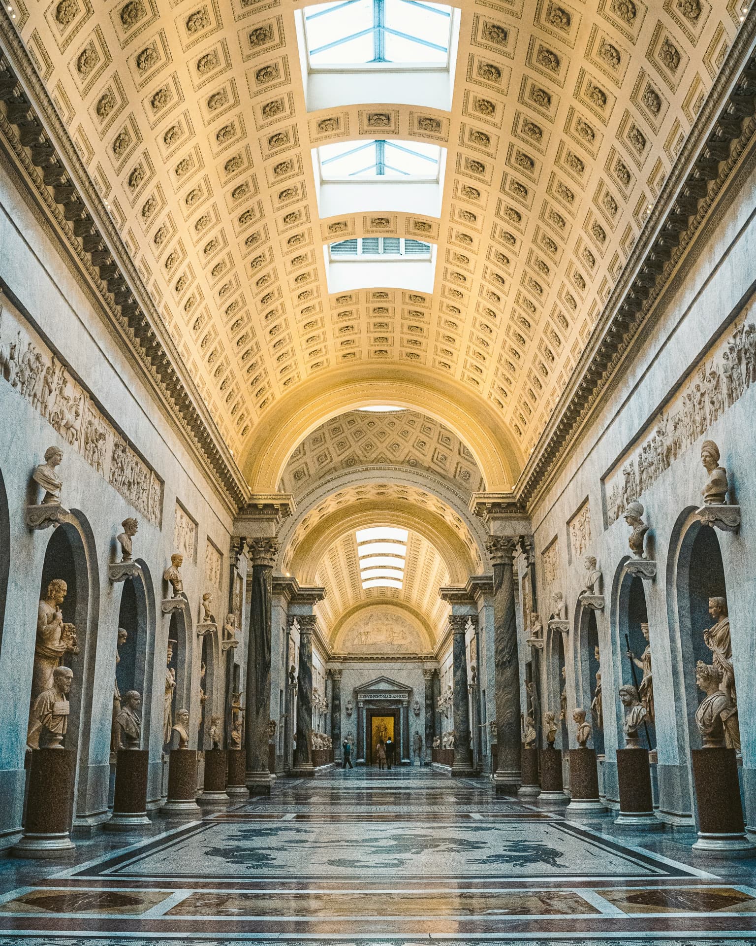 A long gallery lined with marble statues under a coffered, skylit vault
