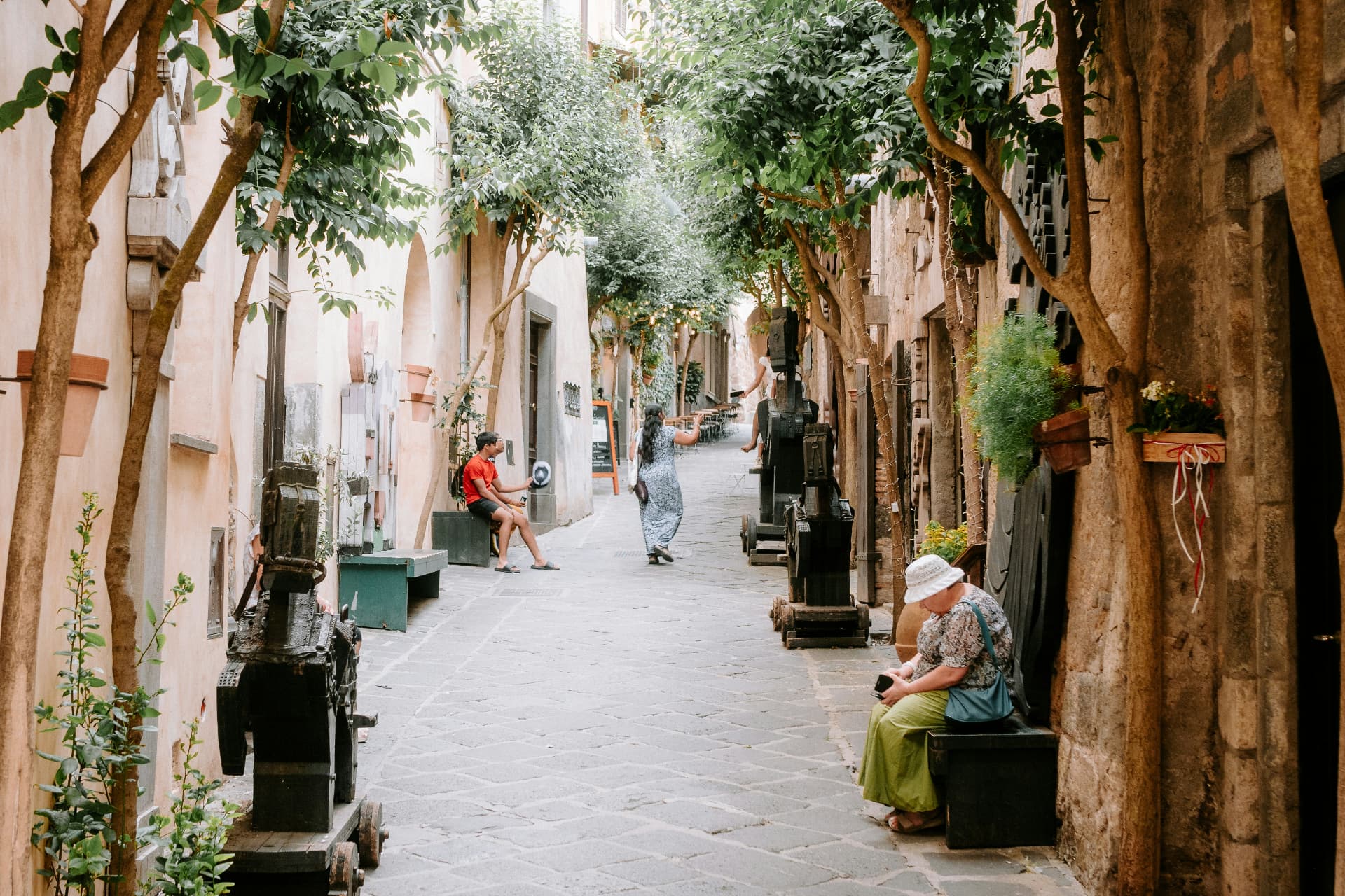 A shaded stone alley in Orvieto, potted plants and wooden sculptures along the lane