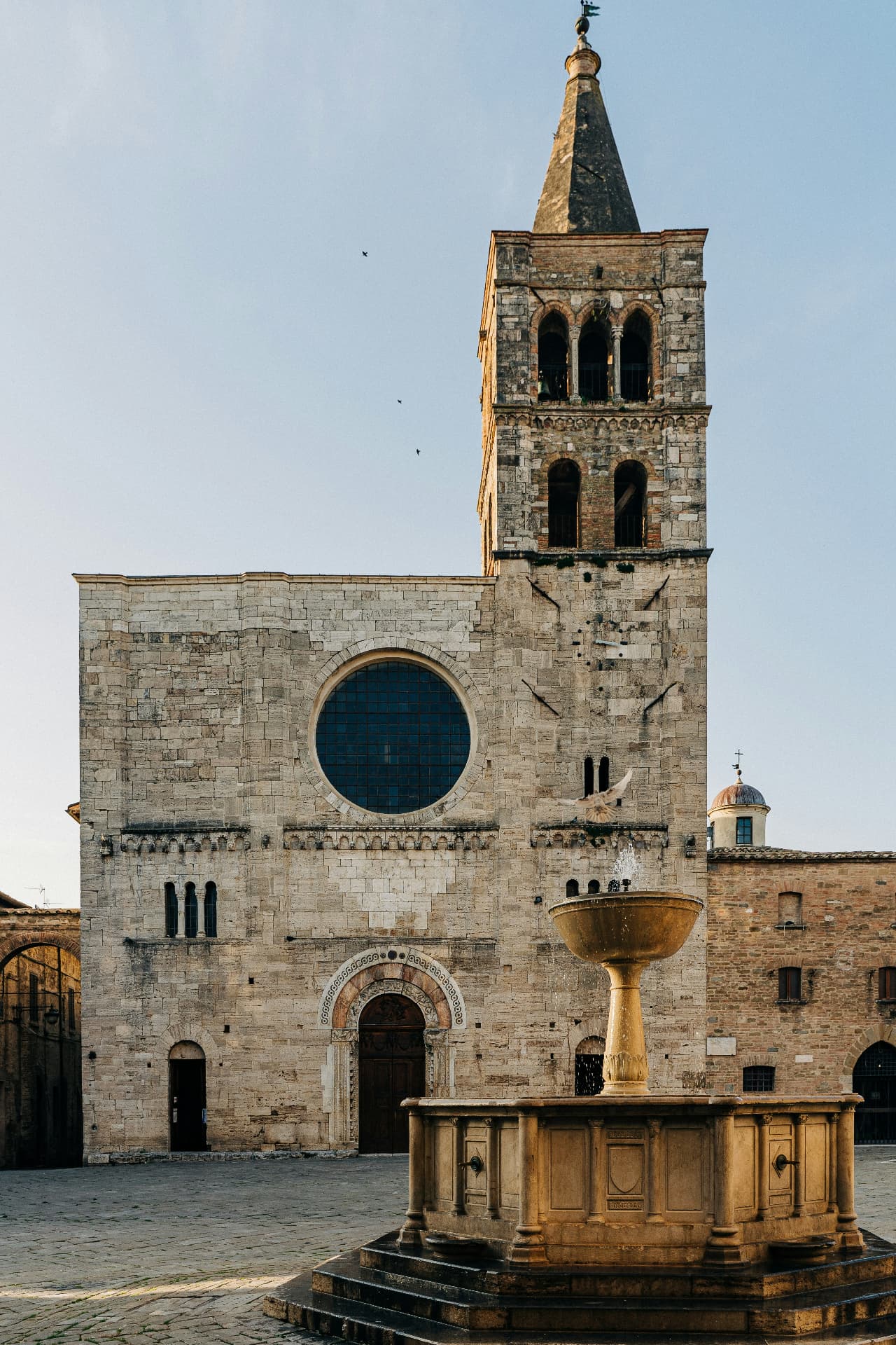 A stone church façade with a round window and an ancient fountain on the piazza