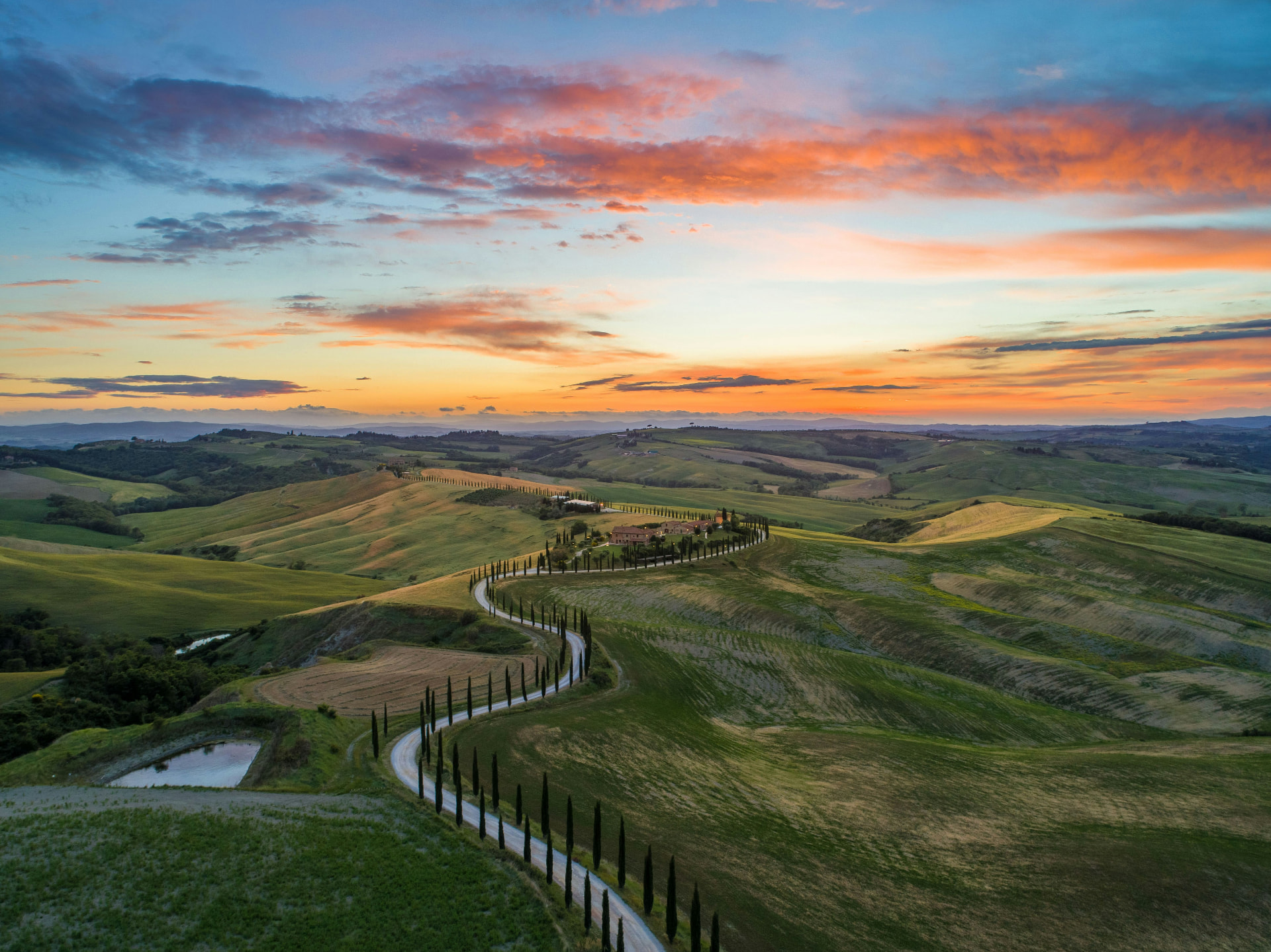 The winding cypress-lined road of the Val d'Orcia under a fiery sunset sky