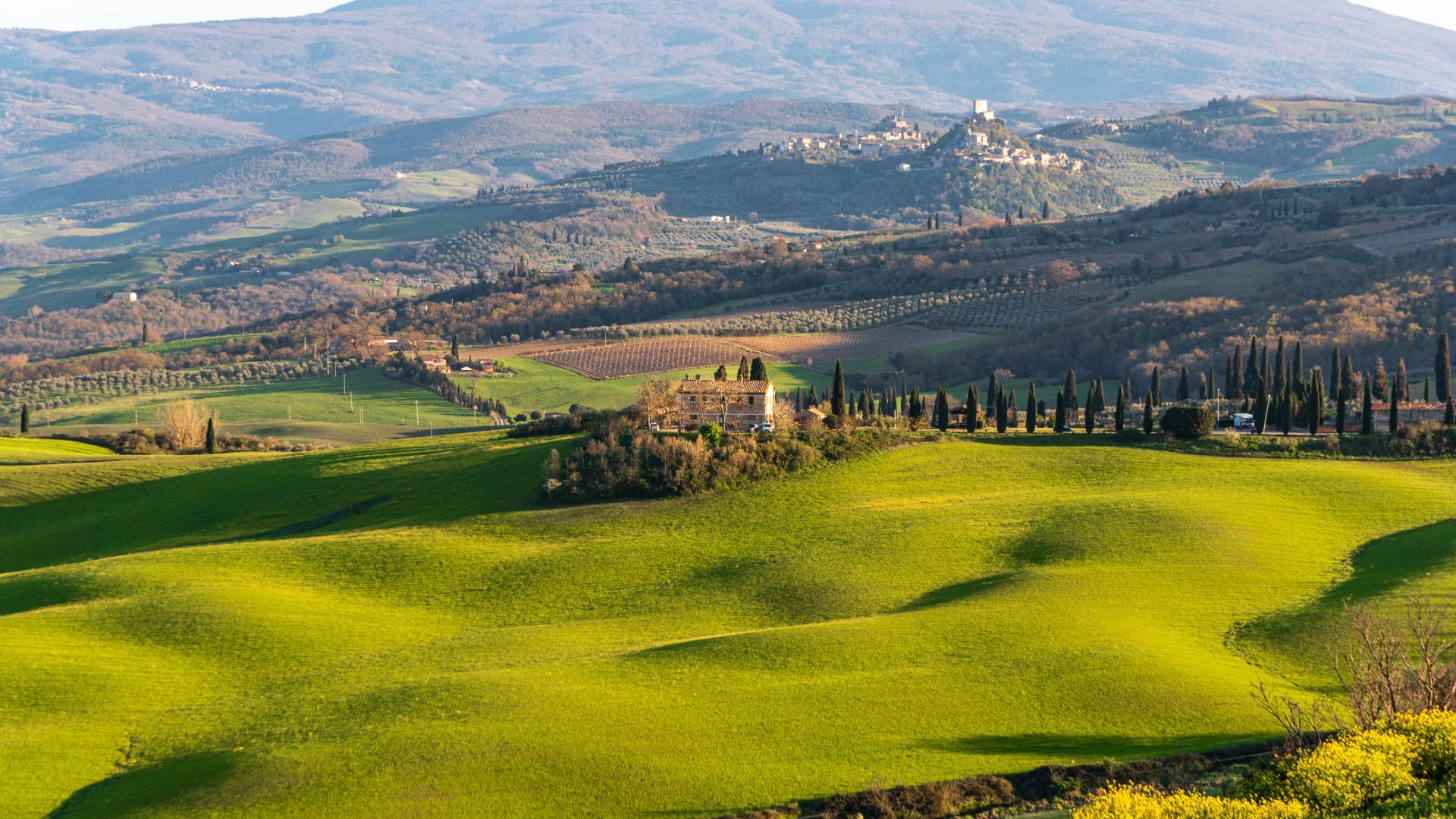 A stone farmhouse among cypress rows and rolling green hills, a hilltop town beyond